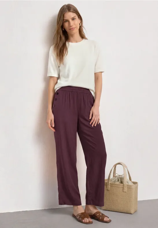 Vrouw in wit T-shirt en bordeauxrode broek, sandalen, met een rieten tas ernaast.