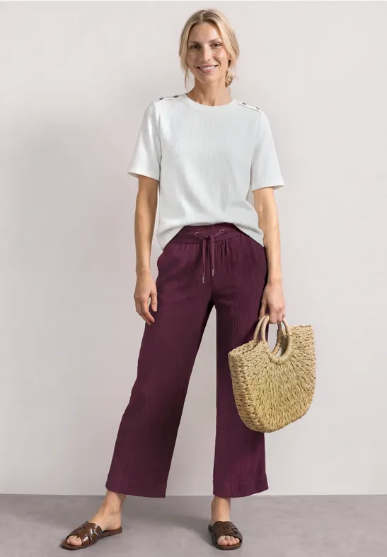 Persoon draagt wit T-shirt, wijde bordeauxrode broek, zwarte sandalen en heeft een gevlochten rieten tas.