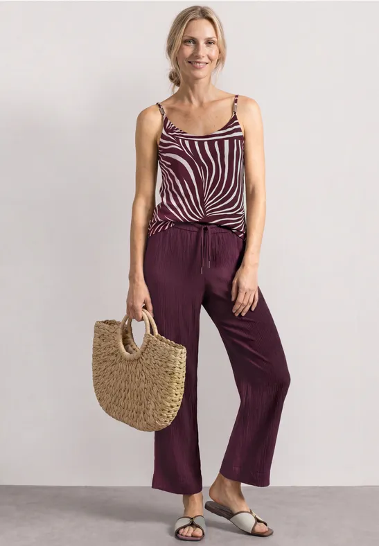 Vrouw met gestreept topje, brede paarse broek, witte sandalen en strooien tas.
