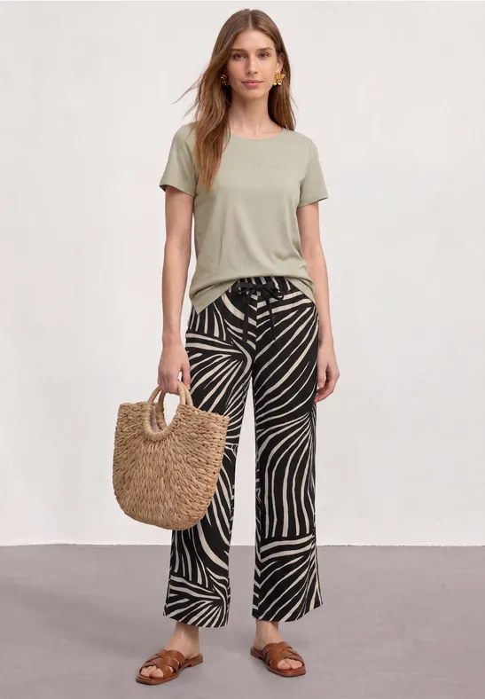 Vrouw met groen T-shirt, wijde zwarte-witte zebradessin broek, sandalen en strooien tas tegen neutrale achtergrond.