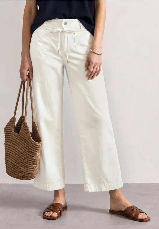 Vrouw in witte, wijde culotte, bruine sandalen, zwarte top en gevlochten strooien tas.
