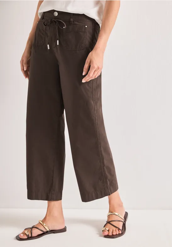 Wijde, enkelhoge broek in donkerbruin met taillekoord, gecombineerd met platte sandaaltjes met smalle bandjes.
