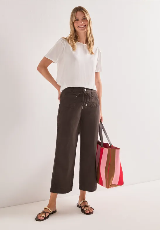 Vrouw in wit T-shirt, wijde donkere broek, zwarte sandalen, met gestreepte boodschappentas.