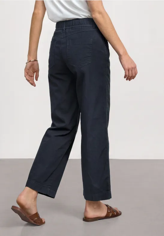 Vrouw van achteren in een wijde donkere jeans en bruine sandalen, neutrale achtergrond.