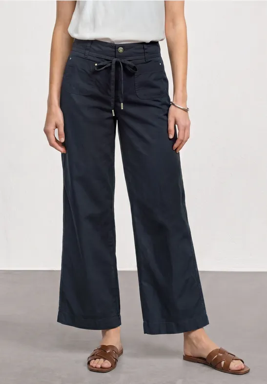 Iemand draagt een wit topje, donkerblauwe, wijde, enkellange broek met tunnelkoord en bruine sandalen.