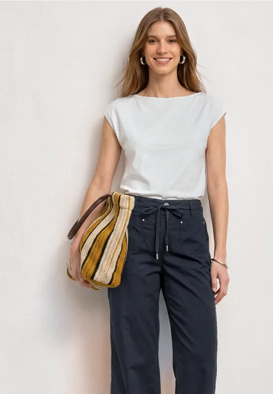 Vrouw met wit hemd, zwarte broek en gestreepte tas, staand voor een lichte achtergrond.