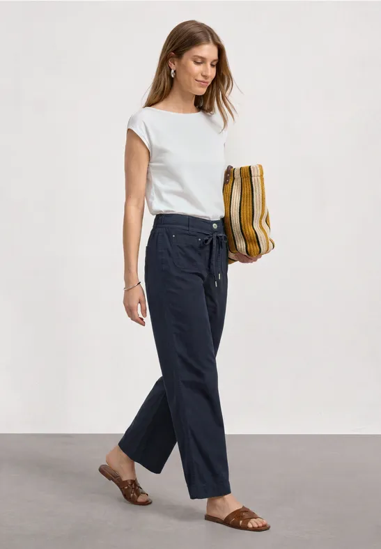 Vrouw met wit t-shirt, losse donkerblauwe broek, platte sandalen en een gestreepte mosterdkleurige tas.