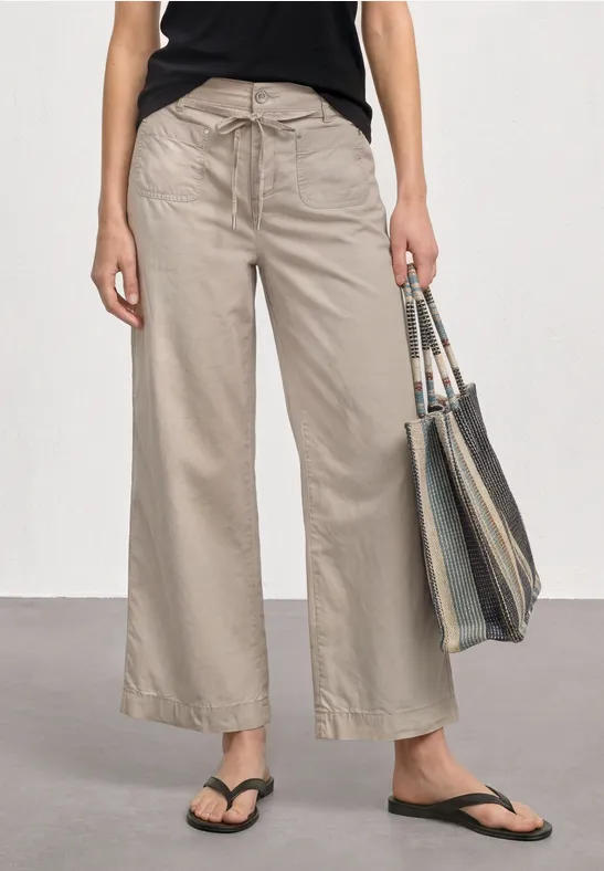 Vrouw draagt losse lichtbeige broek, zwart topje, slippers en houdt een gestreepte tas vast.