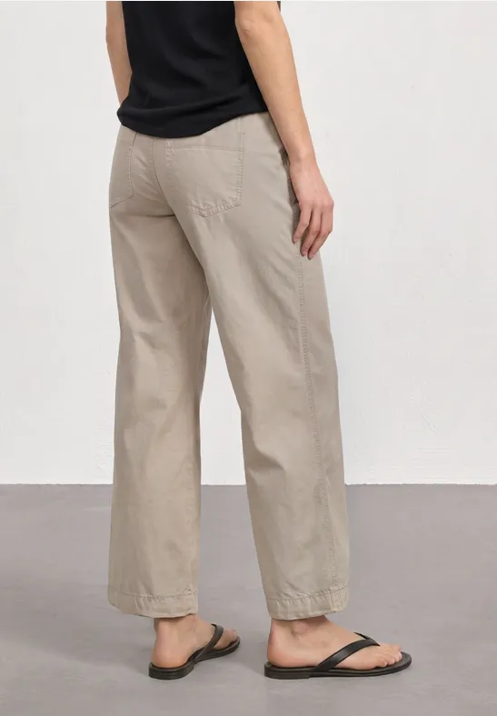 Persoon van achteren in een beige wijde broek, zwart shirt en zwarte sandalen tegen een neutrale achtergrond.