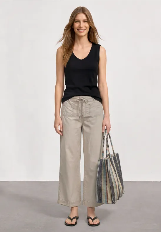 Vrouw in zwart topje, beige wijde broek en zwarte sandalen draagt een gestreepte shopper.