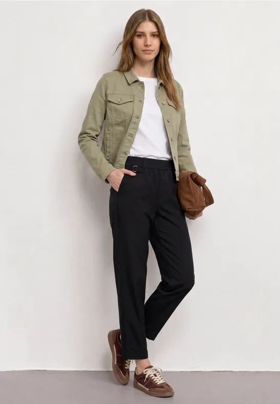 Frau vor weißer Wand, grüne Jacke, weißes Shirt, schwarze Hose, braune Schuhe und Tasche.