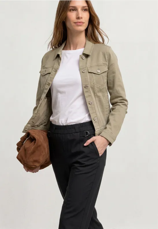 Frau in hellbeiger Jacke, weißem T-Shirt und schwarzen Hosen hält braune Tasche, Hand in der Tasche.