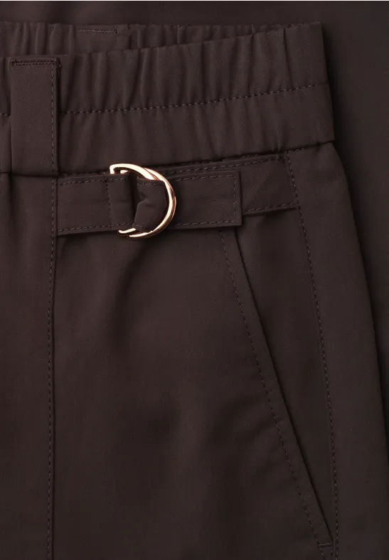 Close-up van een donkerbruine broek met elastische taille, zijzak en een gouden D-ringriem.
