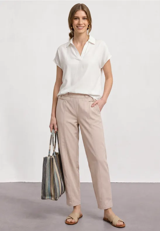 Femme en polo blanc et pantalon beige, sandales, sac rayé, posant devant un fond neutre.