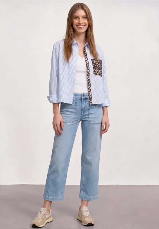 Femme en chemise bleu clair à poche imprimée, top blanc, jean droit bleu et baskets.