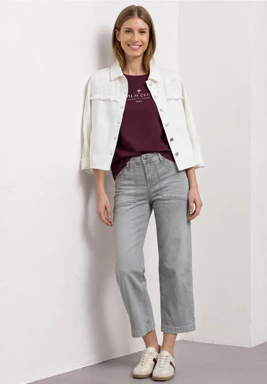 Femme devant un mur blanc, portant une veste blanche, un t-shirt bordeaux, un jean gris droit et des baskets blanches.