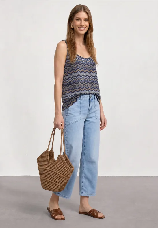 Vrouw met een mouwloos topje met patroon, lichte wijde jeans, bruine sandalen en gevlochten strooien tas.