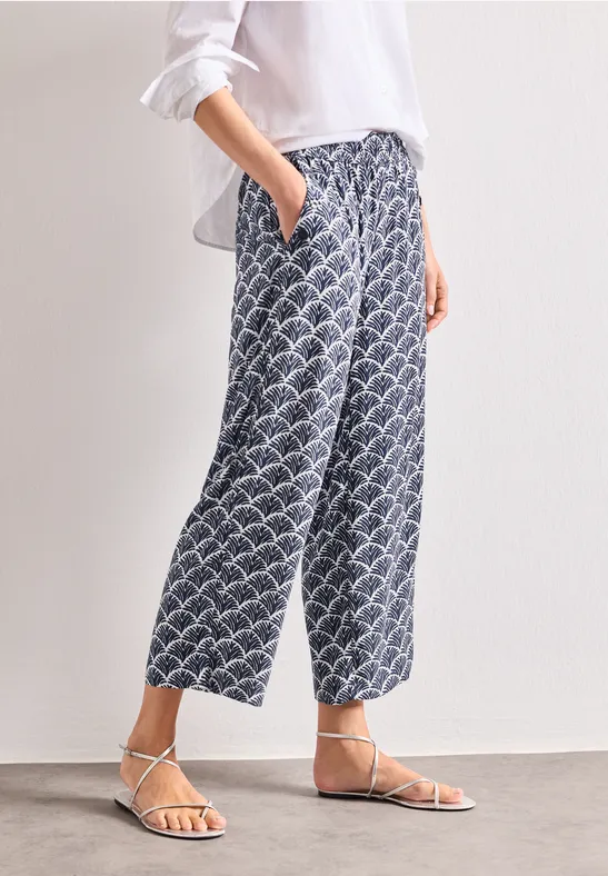 Femme en blouse blanche, pantalon ample bleu-blanc à motifs et sandales plates.