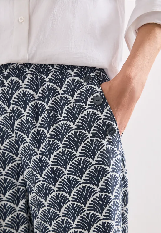 Chemise en lin blanche et pantalon culotte bleu marine à imprimé éventail blanc, main dans la poche.
