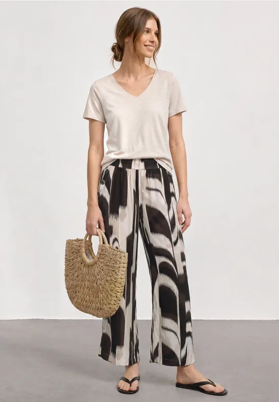 Vrouw in een los wit T-shirt, wijde broek met print, zwarte slippers en strooien tas.