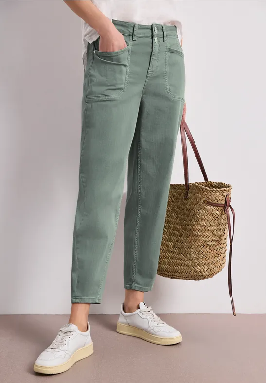 Femme porte un pantalon vert décontracté, baskets blanches et tient un panier tressé.