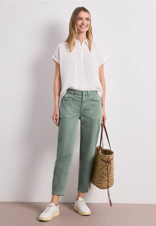 Femme portant chemisier blanc, pantalon vert ample, baskets blanches et sac en osier sur fond clair.
