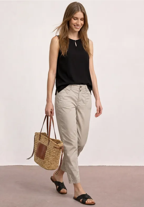 Femme en top noir sans manches, pantalon en lin beige, sandales noires et sac en osier devant un fond neutre.
