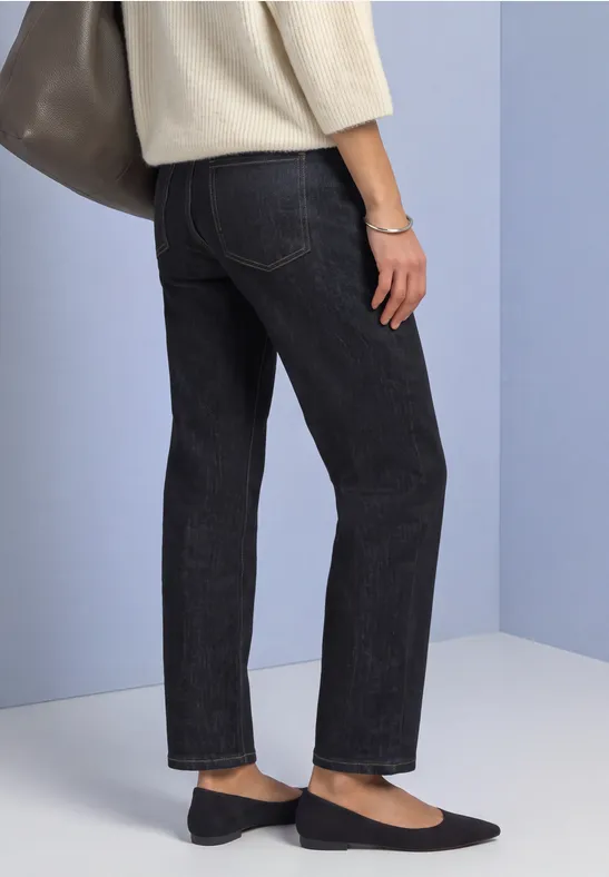 Seitliche Ansicht einer Person in dunkelgrauen Jeans, cremefarbenem Oberteil, schwarzen Ballerinas und Tasche.