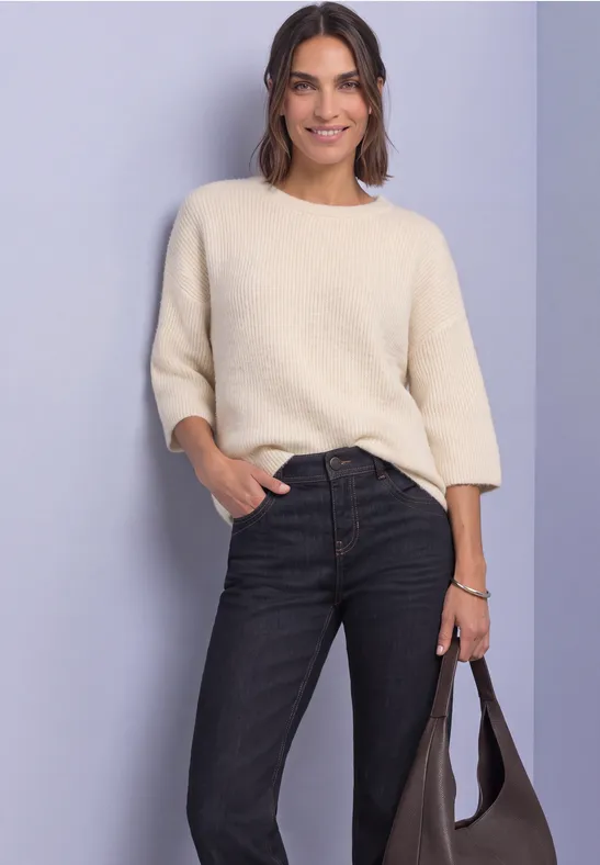 Frau im cremefarbenen Pullover mit 3/4-Ärmeln, schwarzen Jeans und Tasche vor neutralem Hintergrund.