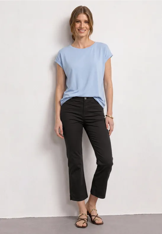 Une personne porte un t-shirt bleu clair, un pantalon 7/8 noir et des sandales plates devant un mur blanc.