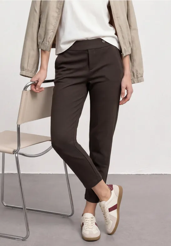 Schmale braune Hose, weiße Sneaker mit beiger Sohle, helle Jacke, sitzend an einem Stuhl.