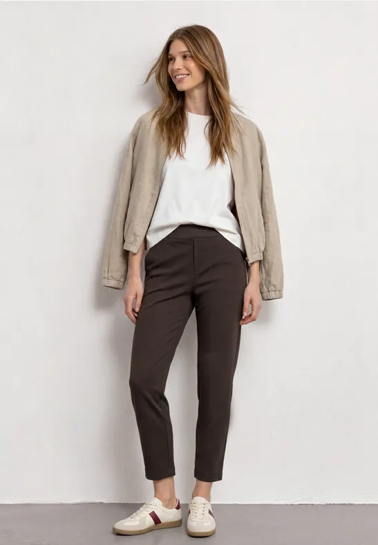 Frau mit beigem Blazer über weißem Shirt, dunklen Hosen und weißen Sneakern, lächelnd in lässiger Pose.