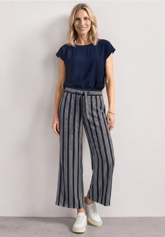 Eine Frau trägt ein dunkelblaues T-Shirt, gestreifte Culotte-Hosen und weiße Sneaker vor hellem Hintergrund.