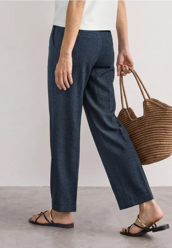 Vrouw in donkerblauwe broek, wit shirt, platte sandalen en met een gevlochten strooien tas.