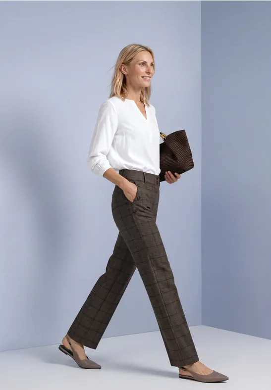 Femme en chemise blanche et pantalon marron à carreaux, marchant avec une pochette marron.