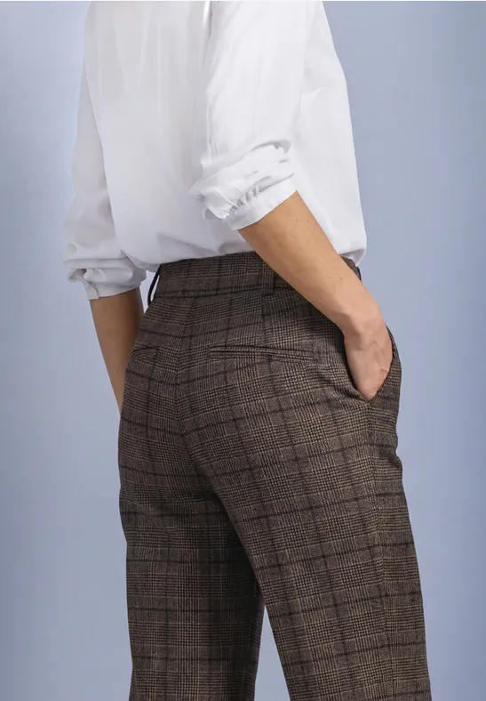 Personne vue de dos en pantalon brun à carreaux et chemise blanche, mains dans les poches.