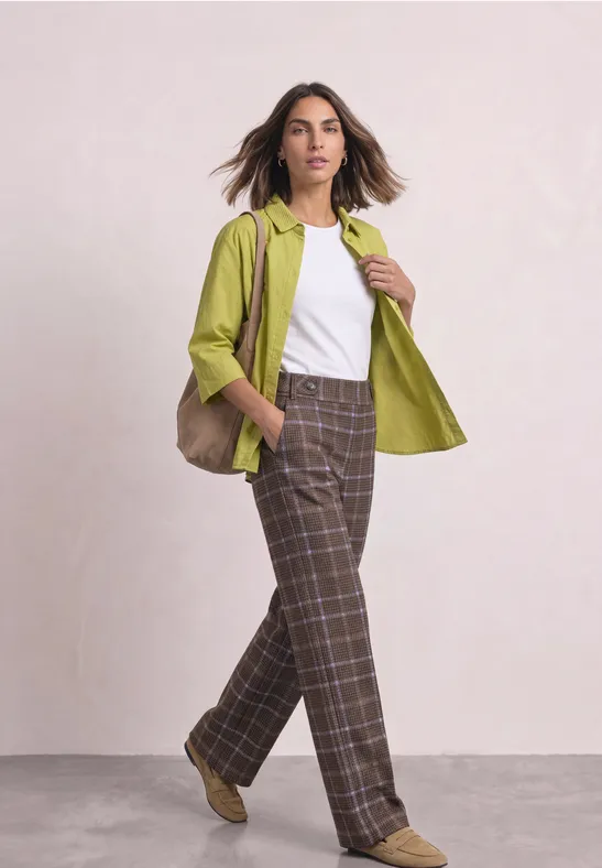 Femme posant devant un mur clair, portant un top blanc, une veste verte et un pantalon à carreaux marron.