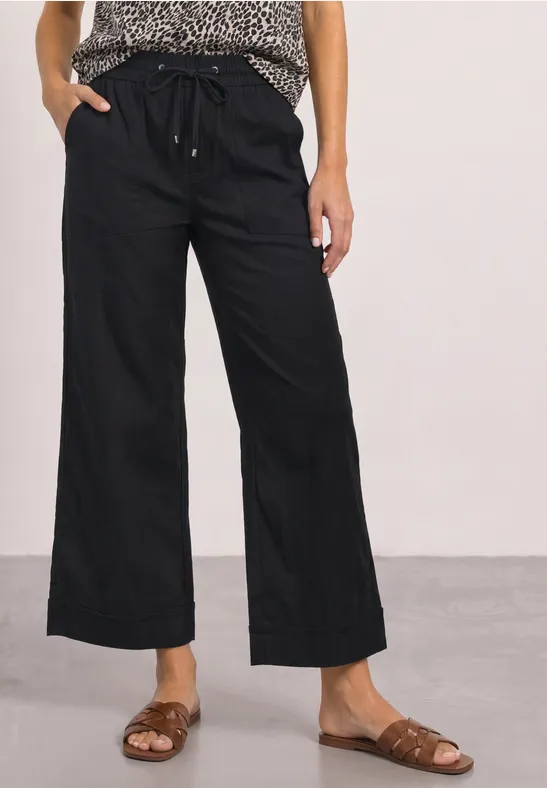 Frau in weiter schwarzer Leinenhose mit Kordelzug und braunen Sandalen, dazu gemustertes Top.