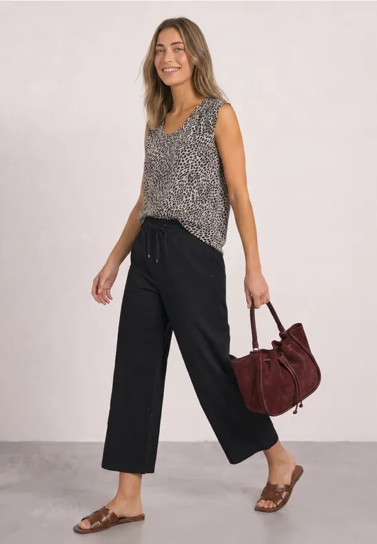 Junge Frau in gemustertem Top, schwarzen Culottes und Sandalen, trägt eine bordeauxrote Tasche.