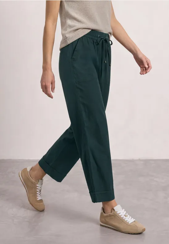 Personne portant un pantalon vert ample à cordon et des baskets beiges, marchant sur un sol gris clair.