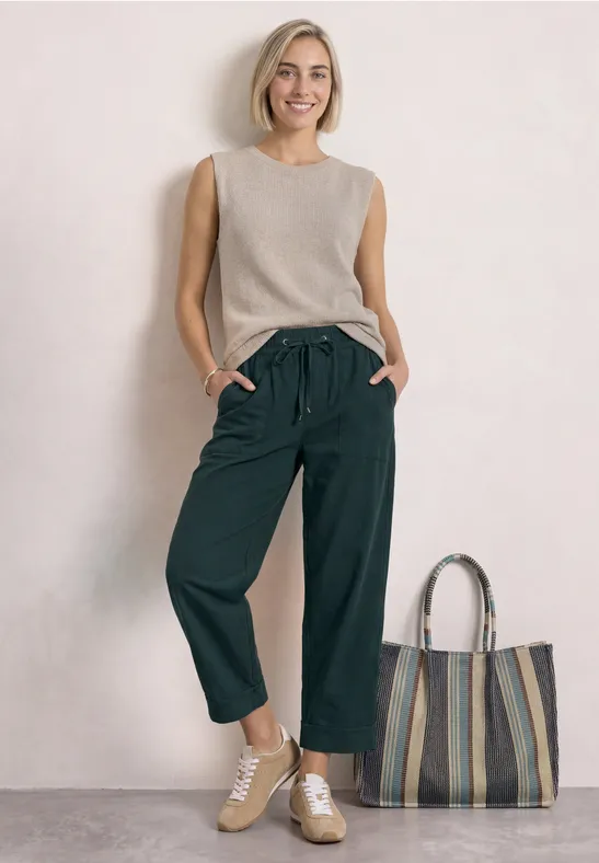 Femme souriante en débardeur beige, pantalon large noir, baskets et grand cabas rayé sur fond clair.