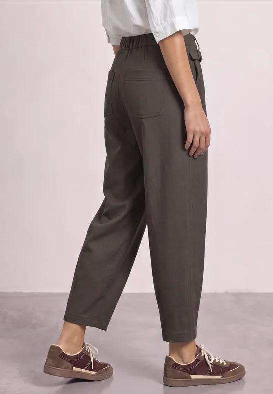 Pantalon gris taille haute, coupe ample et chevilles dégagées, porté avec des baskets marron et un haut blanc.