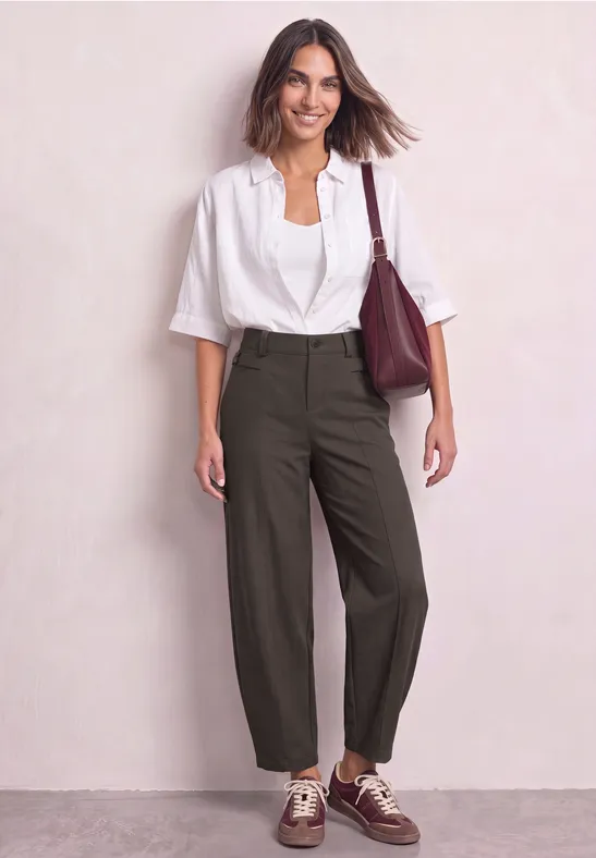 Une femme souriante devant un fond clair porte une blouse blanche, un pantalon ample sombre, des chaussures marron et un sac bordeaux.
