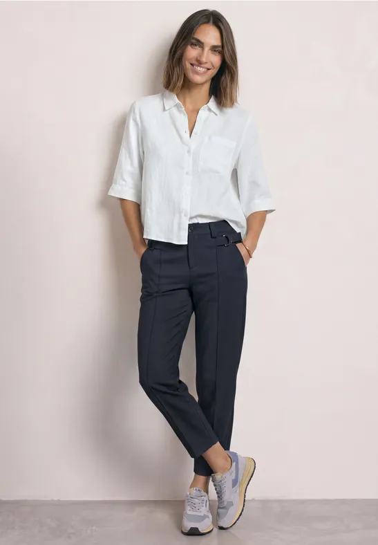 Femme souriante en chemise blanche, pantalon sombre et baskets sur fond clair.