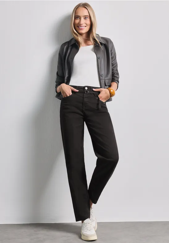 Femme détendue en top blanc, veste et jean noirs, baskets blanches, souriante devant un mur gris.