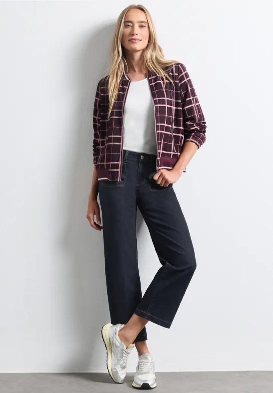 Vrouw in wit topje, bordeaux geruite flanellen blouse, zwarte wijde broek en witte sneakers tegen een witte achtergrond.