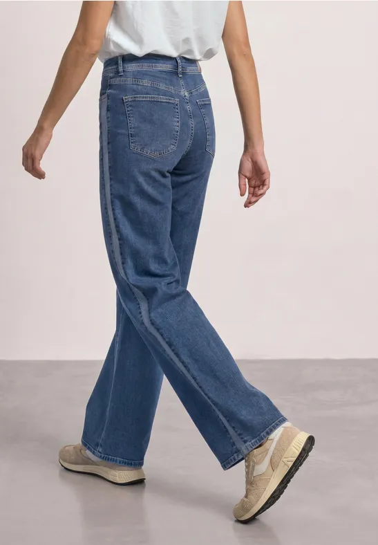 Rückansicht einer Person in weitem blauen Jeans und hellen Schuhen vor neutralem Hintergrund.