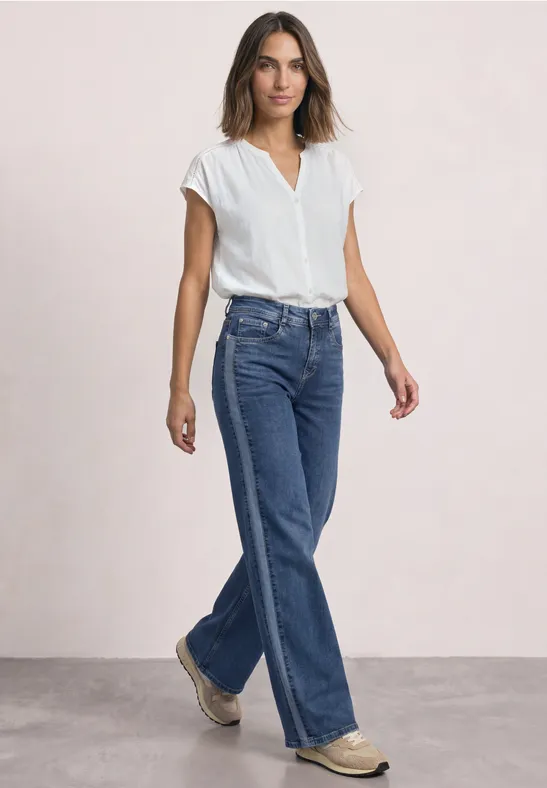 Frau in weißer Bluse und weiten blauen Jeans geht vor hellem Hintergrund.