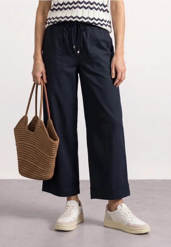 Person trägt dunkelblaue weite Hose, gestreiftes Top, weiße Sneaker und braune geflochtene Tasche.