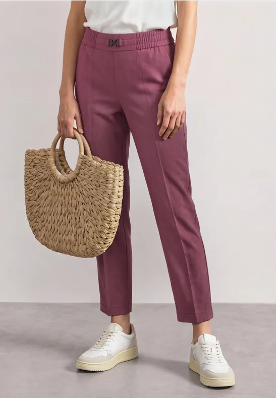 Personne portant un pantalon rose, des baskets blanches et tenant un sac en paille tressée.